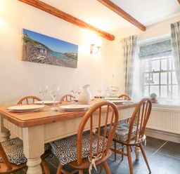 A dining room with a wooden table set with plates glasses and a pitcher surrounded by wooden chairs with cushions and a window with curtains at Aunt Doras in Port Isaac