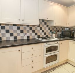A kitchen with upper and lower cabinets black countertop a gas stove double oven microwave and checkered tile backsplash at Ty Potiwr in Conwy