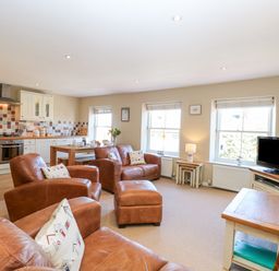 An open-plan living room with brown leather chairs and sofa a kitchen with white cabinets and tiled backsplash and a dining table at Crab Corner in Wells-Next-The-Sea