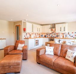 An open plan living area with brown leather sofas and armchair a kitchen with white cabinets and a wooden dining table with breakfast items at Crab Corner in Wells-Next-The-Sea