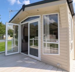 An exterior view of a light beige mobile home with white framed glass doors and windows on a wooden deck at 7 The Oaks in Long Preston near Hellifield