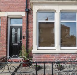 Front view of a brick house with a black door numbered 18 two white framed windows and several potted plants behind a decorative metal fence at 18 Acklington Road in Amble