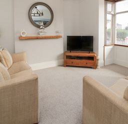 A living room with two beige sofas a wall mirror a wooden shelf with decorations a television on a wooden stand and windows with blinds at 18 Acklington Road in Amble