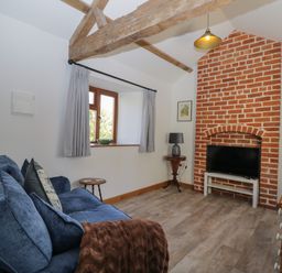 A living room with a blue sofa a television on a white stand in a brick fireplace alcove exposed wooden beams and a window with curtains at Keepers Barn Bromyard