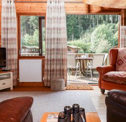 A living room with leather and patterned armchairs a tv on a stand and a sliding door opening to a deck with patio furniture at Trout River Retreat Clifford Bridge near Moretonhampstead