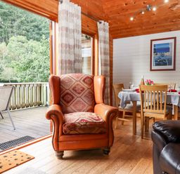 A wooden interior room with an armchair near a dining table set for four and a deck with outdoor chairs at Trout River Retreat Clifford Bridge near Moretonhampstead