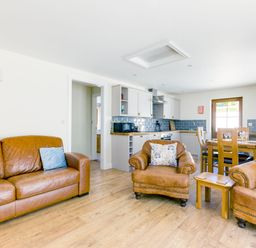 A living room with brown leather sofas and armchairs next to a kitchen with white cabinets and a wooden dining table at Chy Pol in St Minver