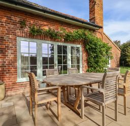 An outdoor patio with a wooden table and six chairs next to a red brick house at Rookery Cottage in Kelsale near Saxmundham