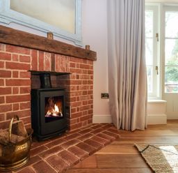 A brick fireplace with a wood burning stove and a brass container with firewood next to a window with curtains and a wooden floor at Rookery Cottage in Kelsale near Saxmundham