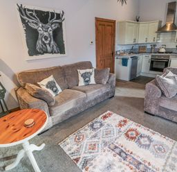 A living room with two sofas and a wooden round table next to a kitchen area with white cabinets at Matfen in Longframlington