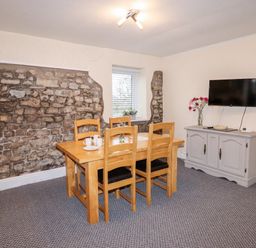 A dining room with a wooden table and chairs a stone accent wall a window a wall mounted TV and a cabinet with flowers at Fern Cottage in St Brides-Super-Ely