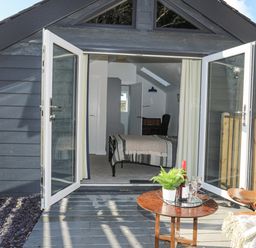 A small house with open glass doors showing a bedroom inside and a wooden patio with a table and chairs at 4 Ty Person in Braichmelyn near Bethesda