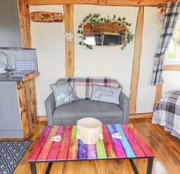 A small living area with a grey sofa, colorful wooden coffee table, kitchenette, and a bed with plaid bedding at Moel Y Parc in Bryniau Clwyd near Denbigh