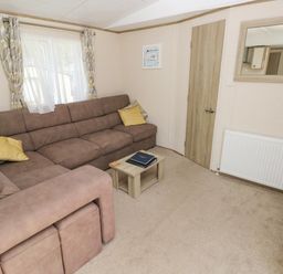 A living room with a brown sectional sofa yellow pillows a small wooden coffee table a window with curtains a door a radiator and a wall mirror at 116 Woodpecker Lodge in Gilfachrheda near Llanarth