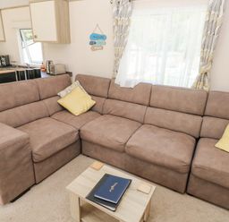 A living room with a brown sectional sofa yellow pillows a small wooden coffee table and a kitchen area at 116 Woodpecker Lodge in Gilfachrheda near Llanarth
