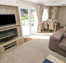 A living room with a brown sofa a television above an electric fireplace and a dining table with chairs at 116 Woodpecker Lodge in Gilfachrheda near Llanarth