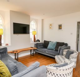 A living room with two grey sofas two patterned chairs a wooden bench with lamps under windows and a TV on the wall at Upper Vestry in Llangrannog