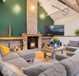 A living room with a stone fireplace a wood burning stove a large grey sectional sofa a coffee table with a chess set and flowers and a wall mounted television at The Waterfall Retreat in Orton
