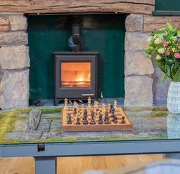 A chessboard on a glass coffee table with a vase of flowers and a wood stove in a stone fireplace at The Waterfall Retreat in Orton