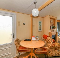 A dining area with a wooden round table and two wicker chairs next to a door and a living room with a floral sofa a fireplace and a television at Cae Gog Near Cemaes Bay