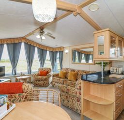 A living room with floral sofas and blue curtains next to a kitchen area with wooden cabinets at Cae Gog Near Cemaes Bay
