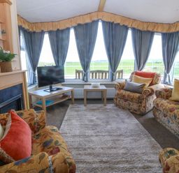 A living room with floral sofas a carpet a television on a stand and large windows with blue curtains at Cae Carreg in Cemaes Bay