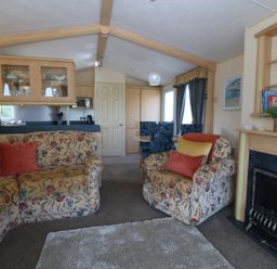 A living room with floral sofas a fireplace a dining table with chairs and kitchen cabinets at Cae Carreg in Cemaes Bay