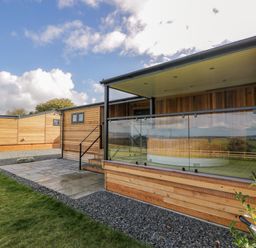 A wooden lodge with a covered porch and glass railing on a stone patio with grass at Betsi Lodge 1 in Llanelidan near Corwen