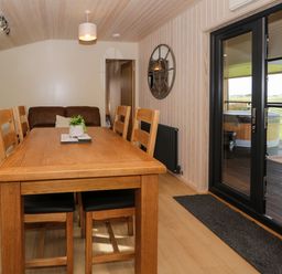 A dining area with a wooden table and six chairs next to black framed glass doors opening to an outdoor deck with a hot tub at Betsi Lodge 1 Llanelidan near Corwen