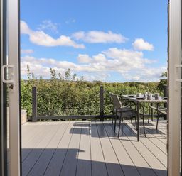 A deck with outdoor table and chairs set for tea with glass railing and hedge in the background at Hazel Lodge in Teigngrace near Newton Abbot