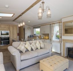 A living room with a sofa, an ottoman, a kitchen area with wooden cabinets, and a wall-mounted electric fireplace at Hazel Lodge in Teigngrace near Newton Abbot