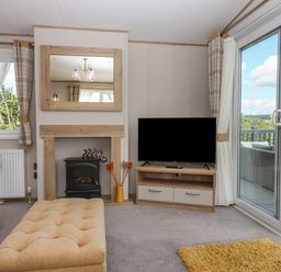 A living room with a TV on a wooden stand a fireplace with a mirror above and sliding glass doors to a deck at Hazel Lodge in Teigngrace near Newton Abbot
