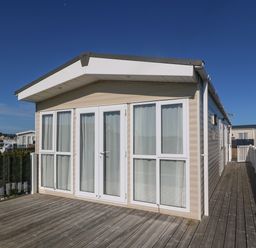 An exterior view of a beige static caravan with large white framed windows and doors on a wooden deck at Seafront Holiday Home 2 in Pwllheli