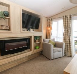 A living room with an armchair a wall-mounted television an electric fireplace and sliding glass doors with curtains at Seafront Holiday Home 2 in Pwllheli