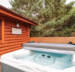 An outdoor hot tub next to a wooden log cabin with a framed sign on the wall at Welcome Hjem Log Cabin Morpeth near Felton