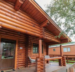 The exterior porch of a wooden log cabin with a door windows chairs and a railing at Welcome Hjem Log Cabin in Morpeth near Felton