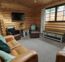 A living room with leather sofas and armchairs a coffee table under a window and a wall-mounted television at Welcome Hjem Log Cabin Morpeth near Felton