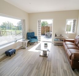 A living room with a brown leather sofa a blue armchair a coffee table a side table with a lamp and books large windows with patterned curtains and a door leading to an outdoor patio at Southend in Welland