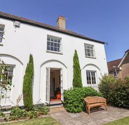 An exterior view of a white two-story house with tall windows and an open door with a wooden bench on a stone patio at Charmouth Lodge in Charmouth