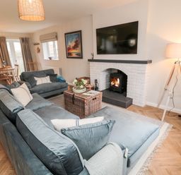 A living room with gray sofas a wicker coffee table a lit fireplace and a dining area at Driftwood Sands in Beadnell