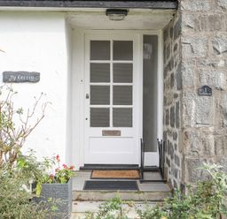A white front door with glass panels and a letterbox between stone walls with plants outside at Ty Cerrig in Llanbedrog