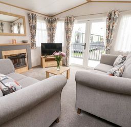 A living room with two gray sofas a wooden coffee table with flowers a fireplace a TV on a stand and large windows with patterned curtains at Silverdale View in East Heslerton near Sherburn