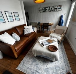 A living room with a brown leather sofa, armchair, ottoman, small dining table with chairs, and beach sign at Waves End in Mevagissey