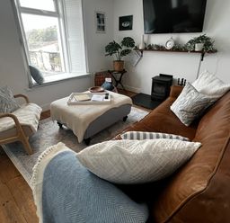 A living room with a brown sofa a wooden chair an ottoman with a tray a window and a wall-mounted television at Waves End Mevagissey