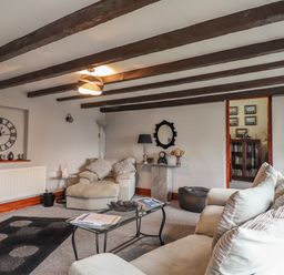 A living room with beams on the ceiling a large clock on the wall a sofa and armchair a glass coffee table and a doorway to another room at High Shaftoe in Ponteland