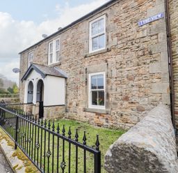 A stone house with white framed windows and a small porch behind a black metal fence at Tyne View Cottage in Bellingham
