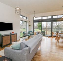 A living room with a gray sofa blue armchairs a wall-mounted TV and a dining table by large windows at Coorie In in Aberfeldy