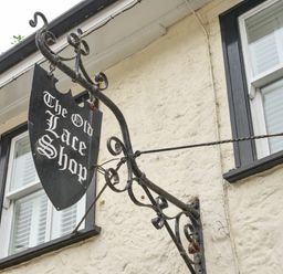A black hanging sign with white text saying The Old Lace Shop on a beige building with two windows at The Old Lace Shop in Beer