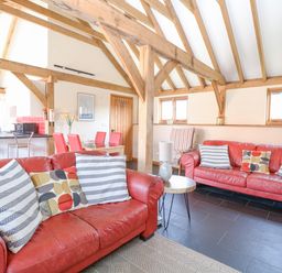 A living room with red sofas, striped and patterned cushions, wooden beams, a small round table with a lamp, and a dining area with red chairs at 5a Hideaways Hunstanton