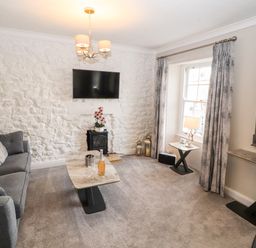 A living room with a grey sofa a marble coffee table a television mounted on a white stone wall and a window with grey curtains at Market Square Maisonette in Kirkby Lonsdale